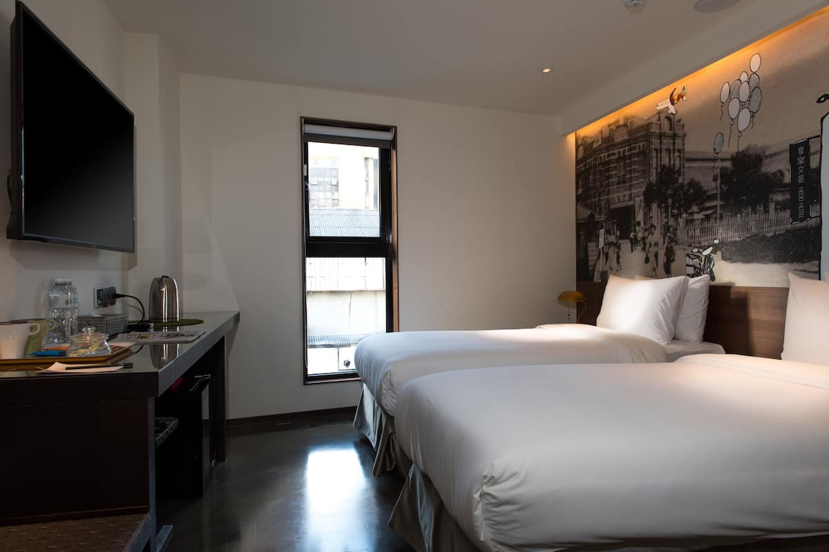 Quarto de hotel com duas camas, uma secretária com chaleira, televisão de tela plana e uma janela grande.