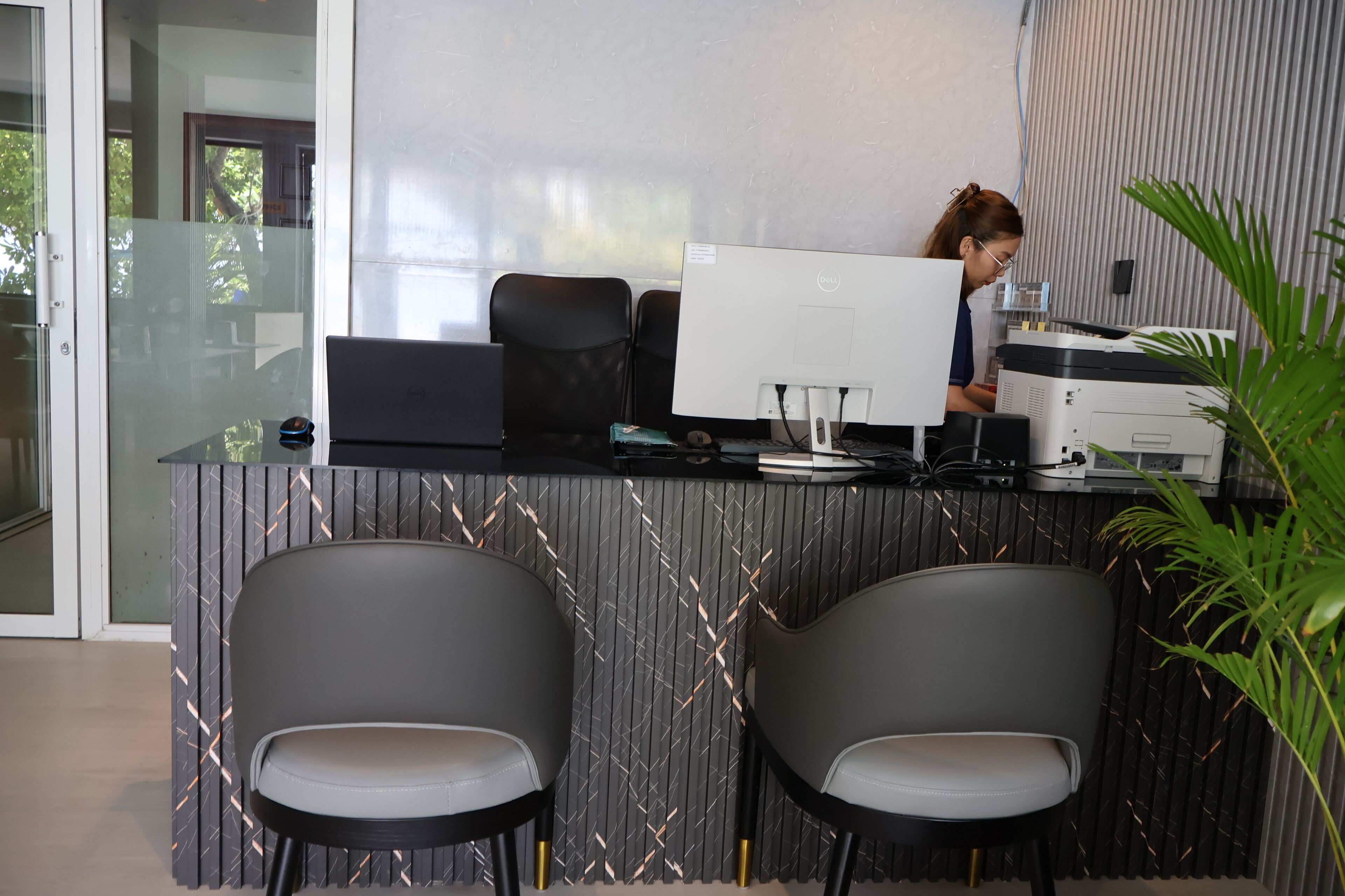 A reception area with a glass partition, two grey chairs, a desk with a computer, and a printer.