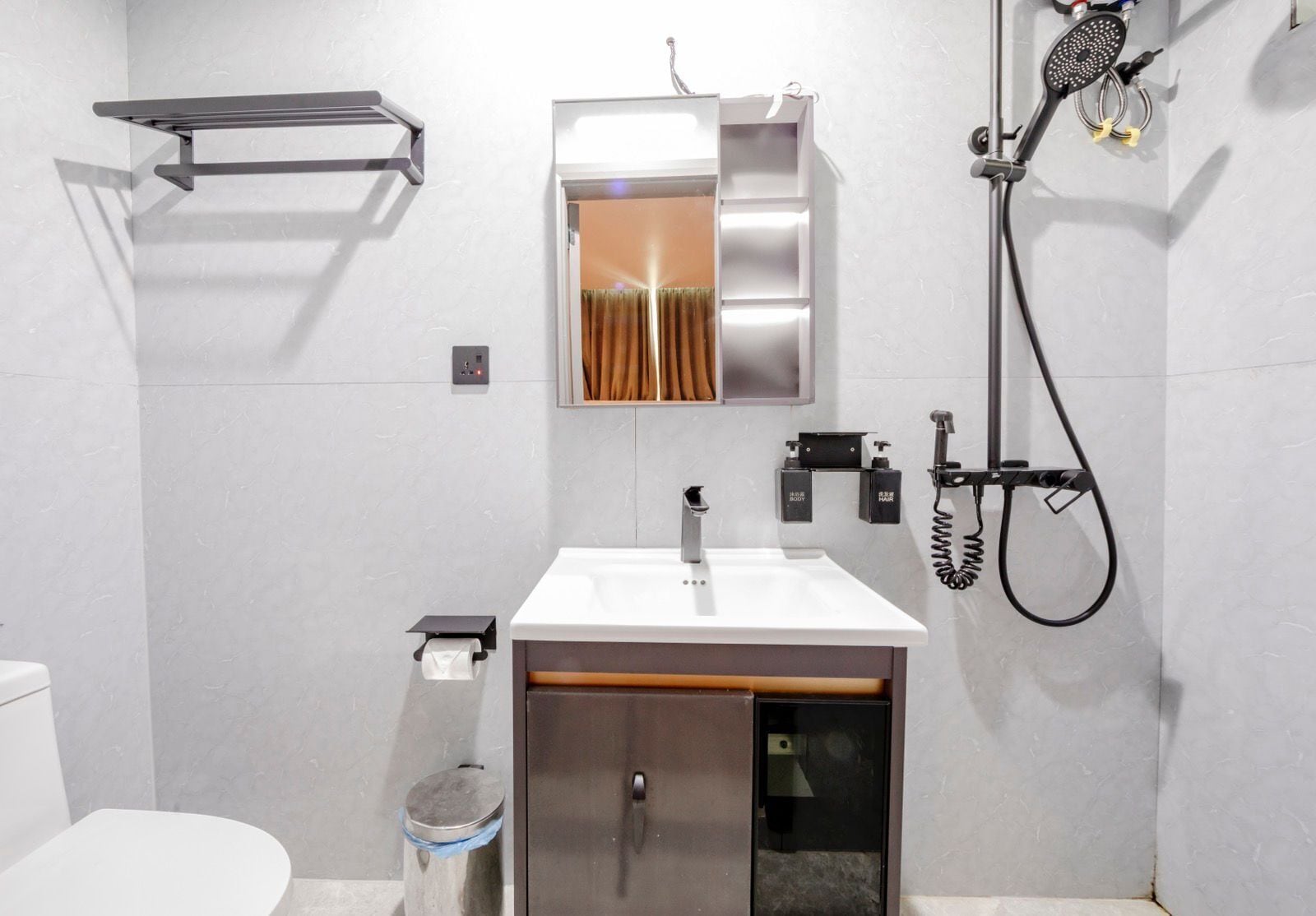 A modern bathroom with a white sink, mirror, and shower. There is a towel rack above the sink and a toilet with a trash can beside it.
