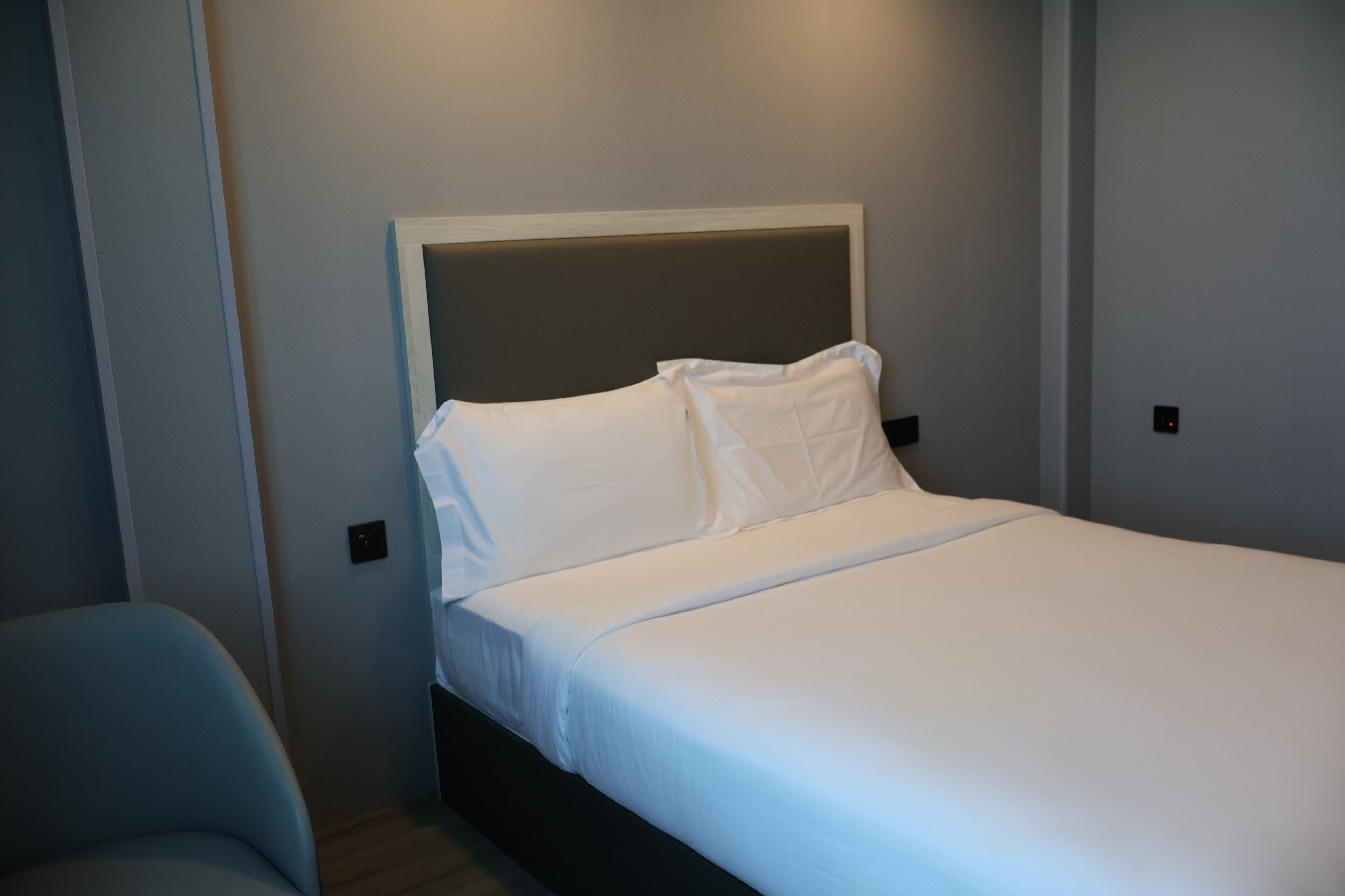 A neatly made bed with white linens in a room with light-colored walls and a dark headboard.