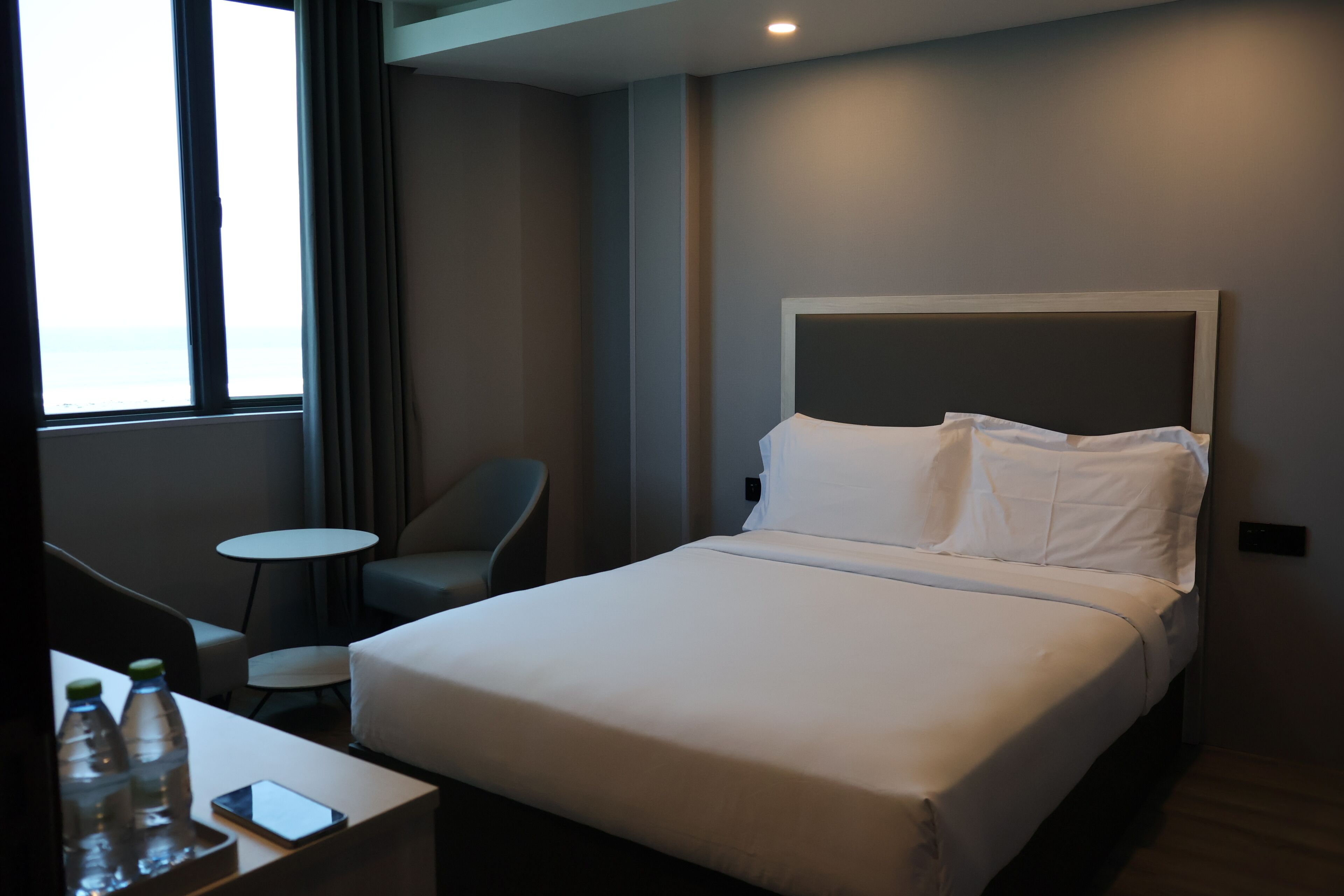 Habitación de hotel con una cama grande, una mesita redonda pequeña, una silla y vista al mar a través de una ventana.