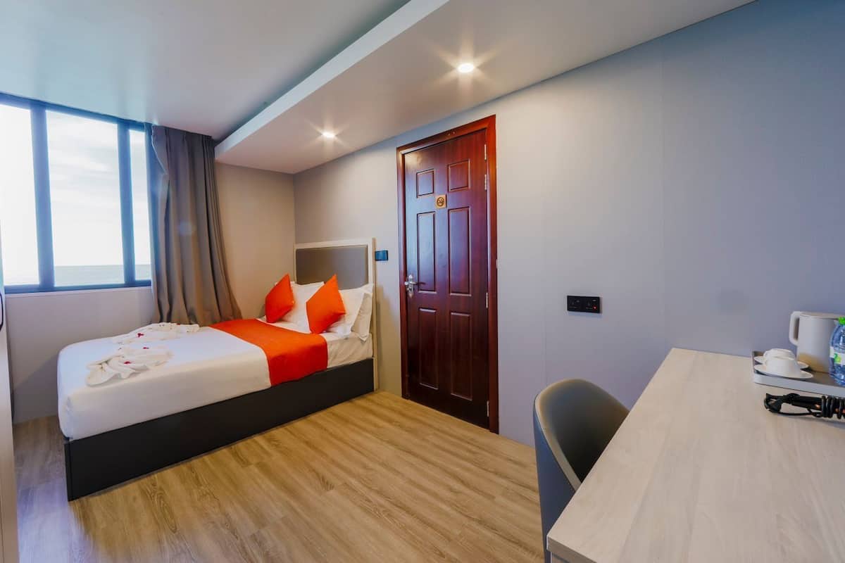 A hotel room with a bed, a desk, a chair, a door, and a window.