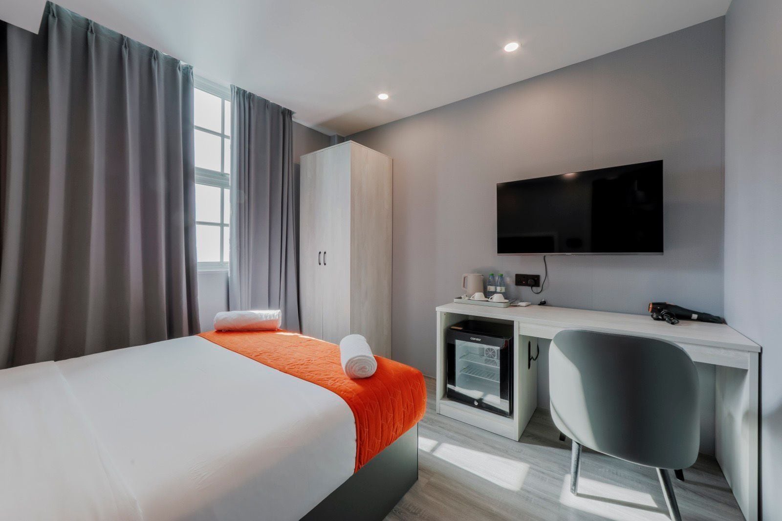 A hotel room with a bed, a desk, a chair, a TV, and a minibar.