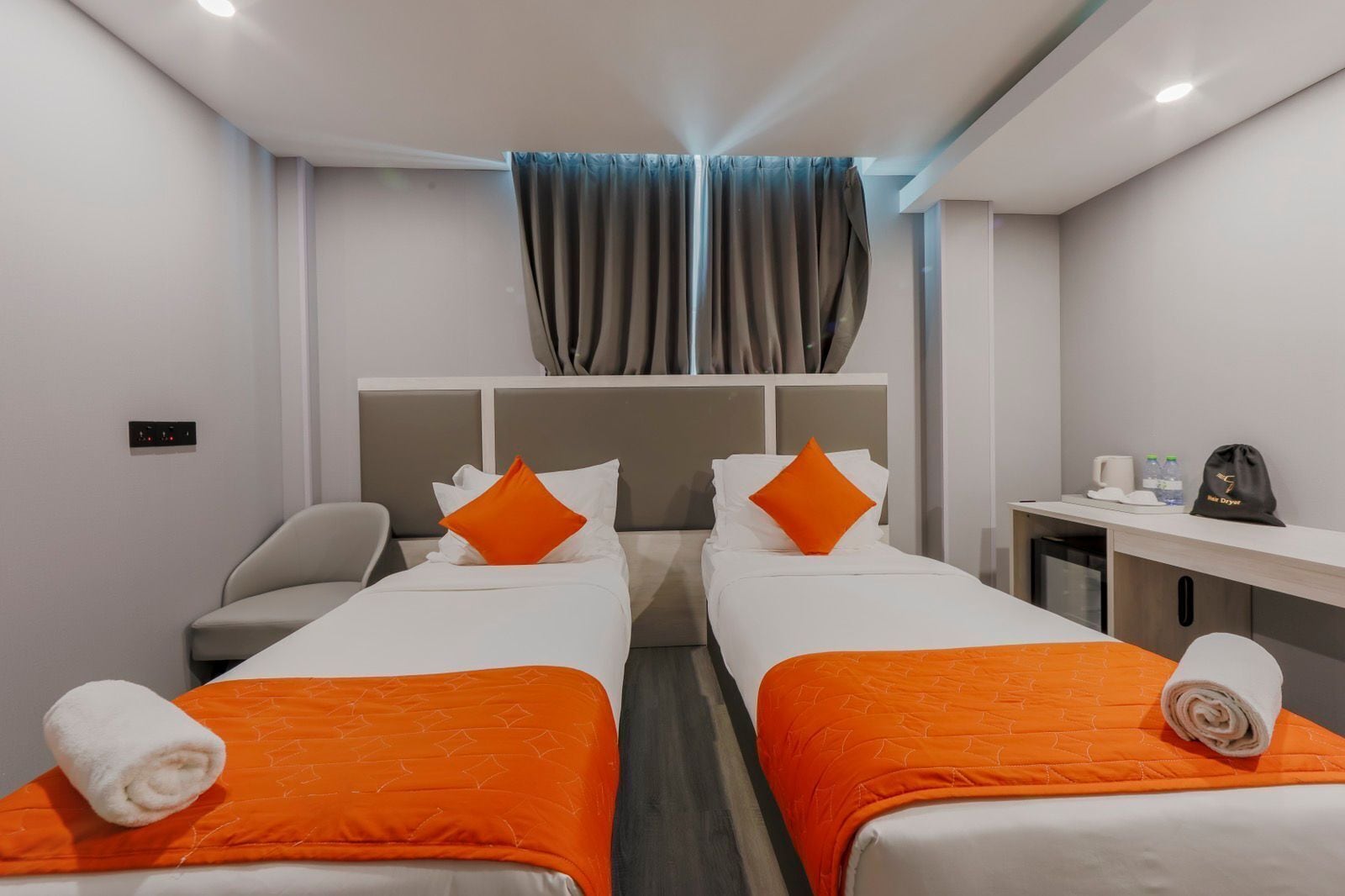 A hotel room with two beds, each with an orange bedspread and pillows, a desk with a chair, a small kitchenette, and a window with curtains.