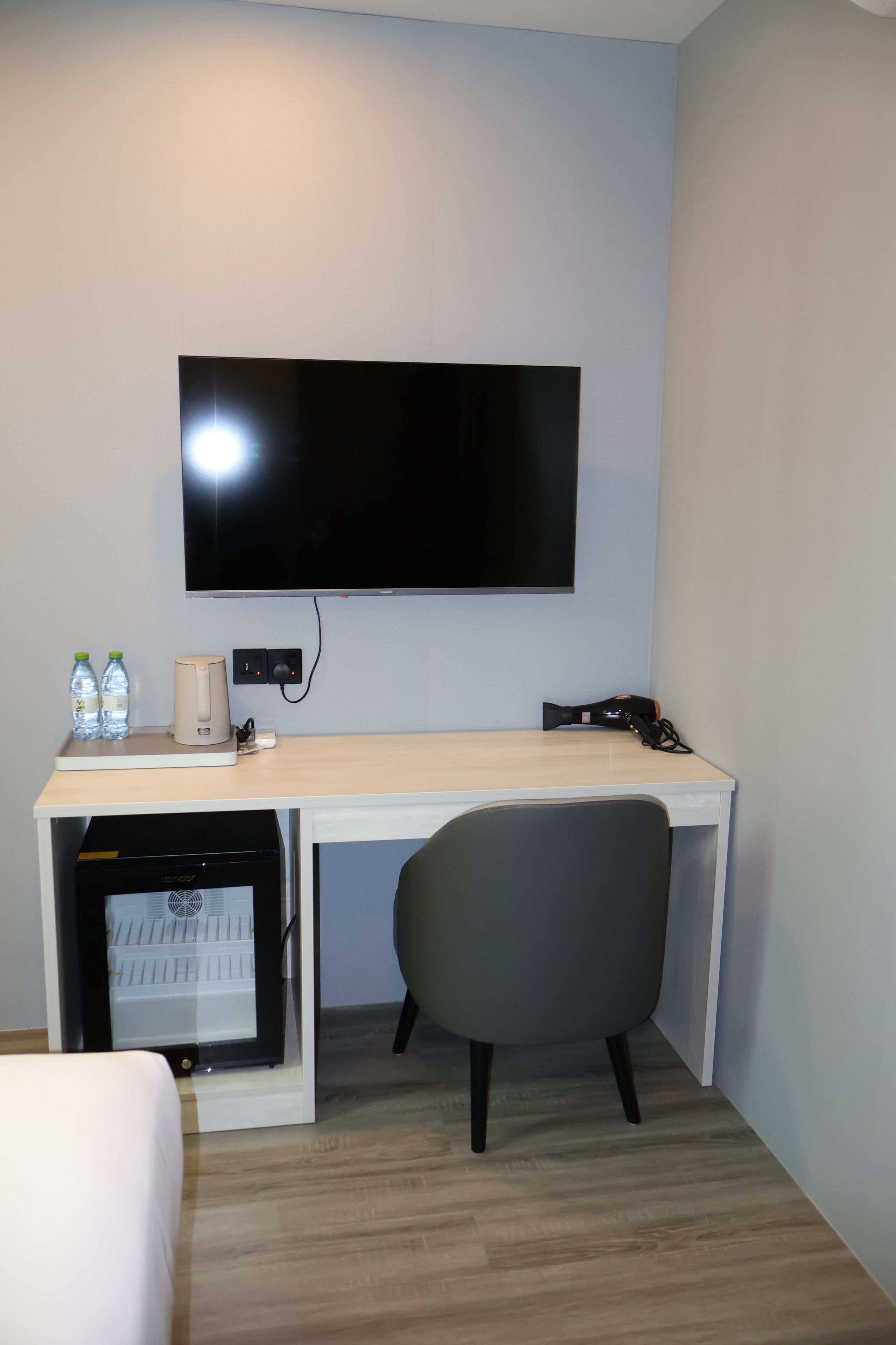 Ein kompaktes Hotelzimmer mit Flachbildfernseher an der Wand, einem kleinen Schreibtisch mit Stuhl, einer Minikühlschrank und zwei Wasserflaschen auf der Ablagefläche.