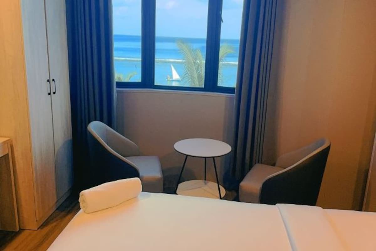 Habitación de hotel con cama, dos sillones, una mesita redonda y vista al océano a través de una ventana.