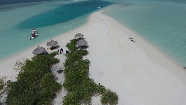 An aerial view of a beach with clear turquoise waters, a boat, and thatched-roof structures.