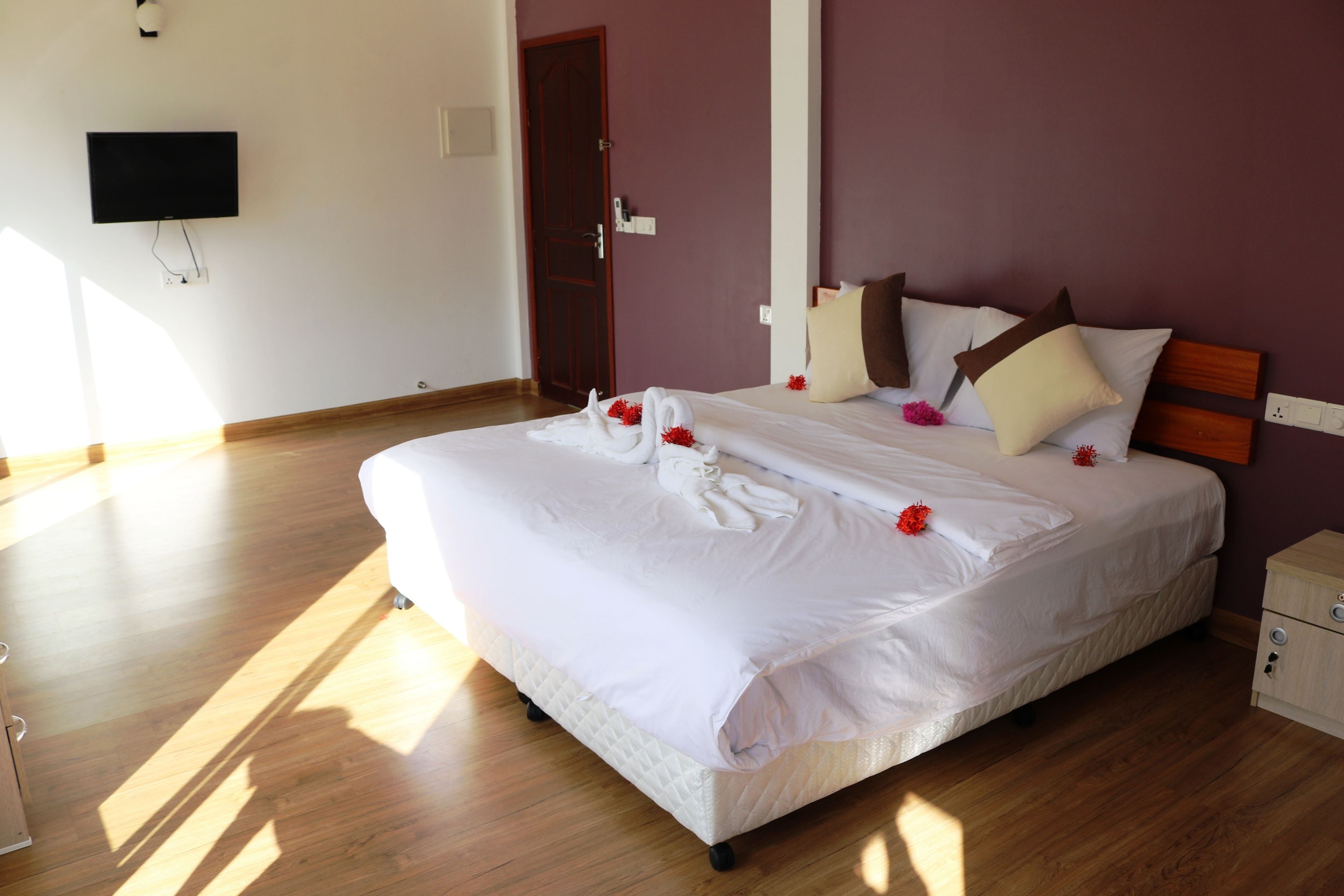 A neatly made bed with white linens and decorative red flowers, a wooden nightstand, and a television mounted on the wall.