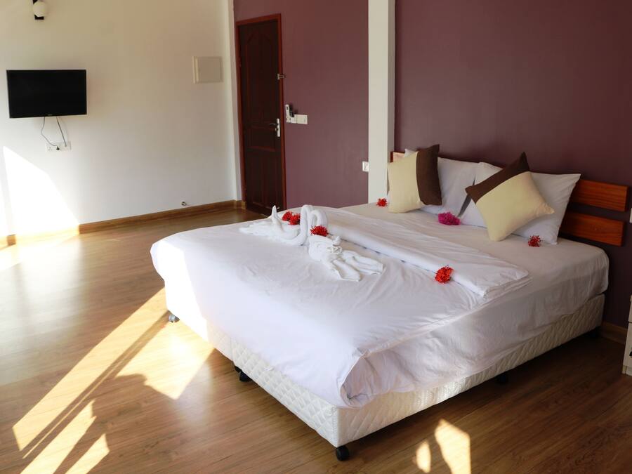 A neatly made bed with white linens and decorative red flowers, a wooden nightstand, and a television mounted on the wall.