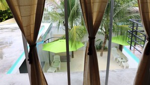 A view through a window with curtains, revealing a patio area with white chairs, a green umbrella, and a staircase.