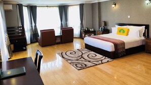A hotel room with a large bed, a desk with a laptop, two armchairs, a patterned rug, and a nightstand with a lamp.