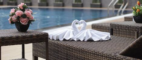 A poolside area with wicker lounge chairs, a heart-shaped towel arrangement, and a vase of pink flowers.
