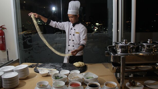 A chef preparing food in a kitchen with a view of the city at night. - Atrium Premiere Hotel Cilacap (Cilacap)