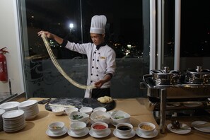 A chef preparing food in a kitchen with a view of the city at night. - Atrium Premiere Hotel Cilacap (Cilacap)