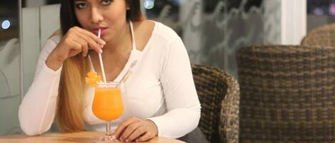 Una mujer sentada a una mesa con una bebida, en una habitación con sillas de mimbre y vista a la ciudad de noche.