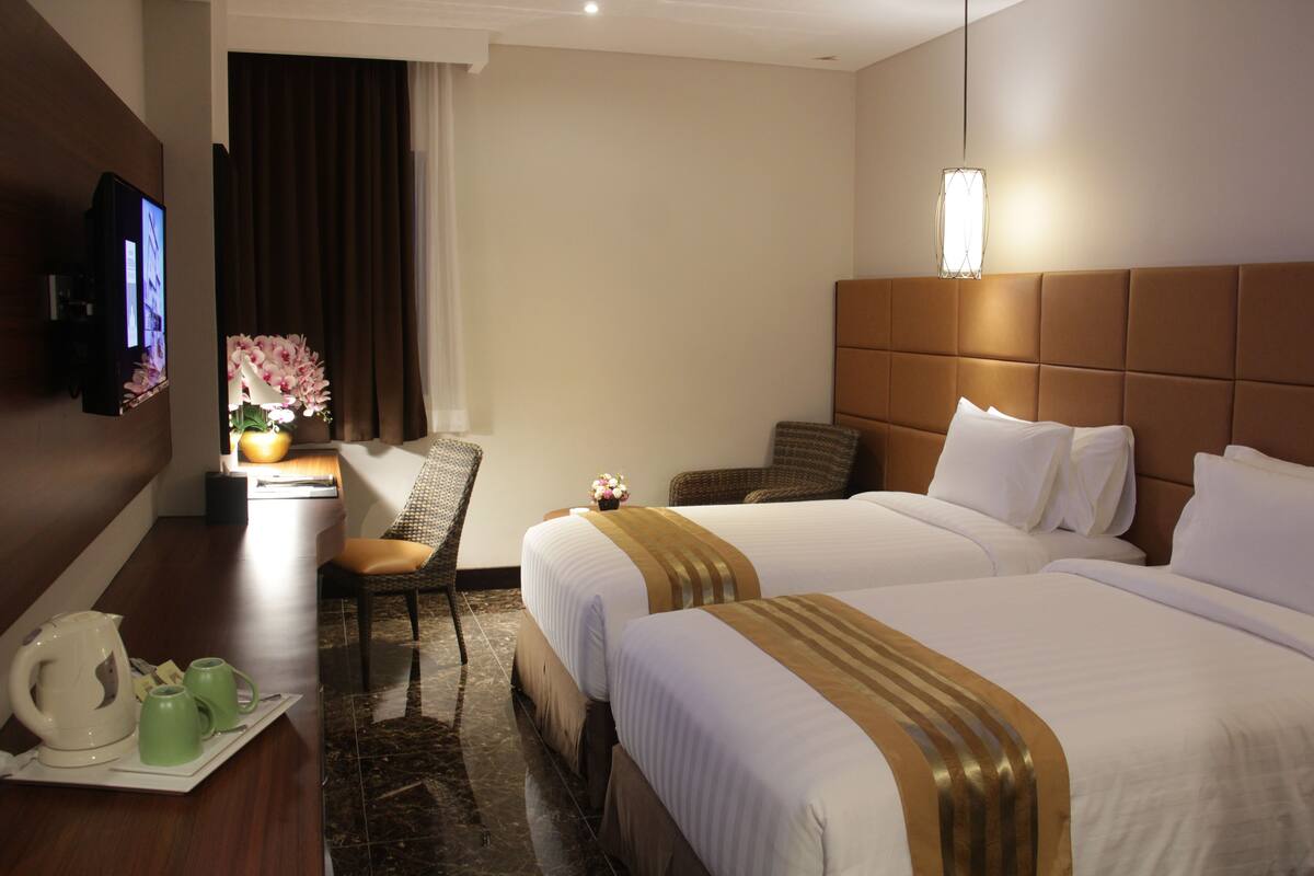 A hotel room with two beds, a TV, a desk with a kettle and cups, and a chair.