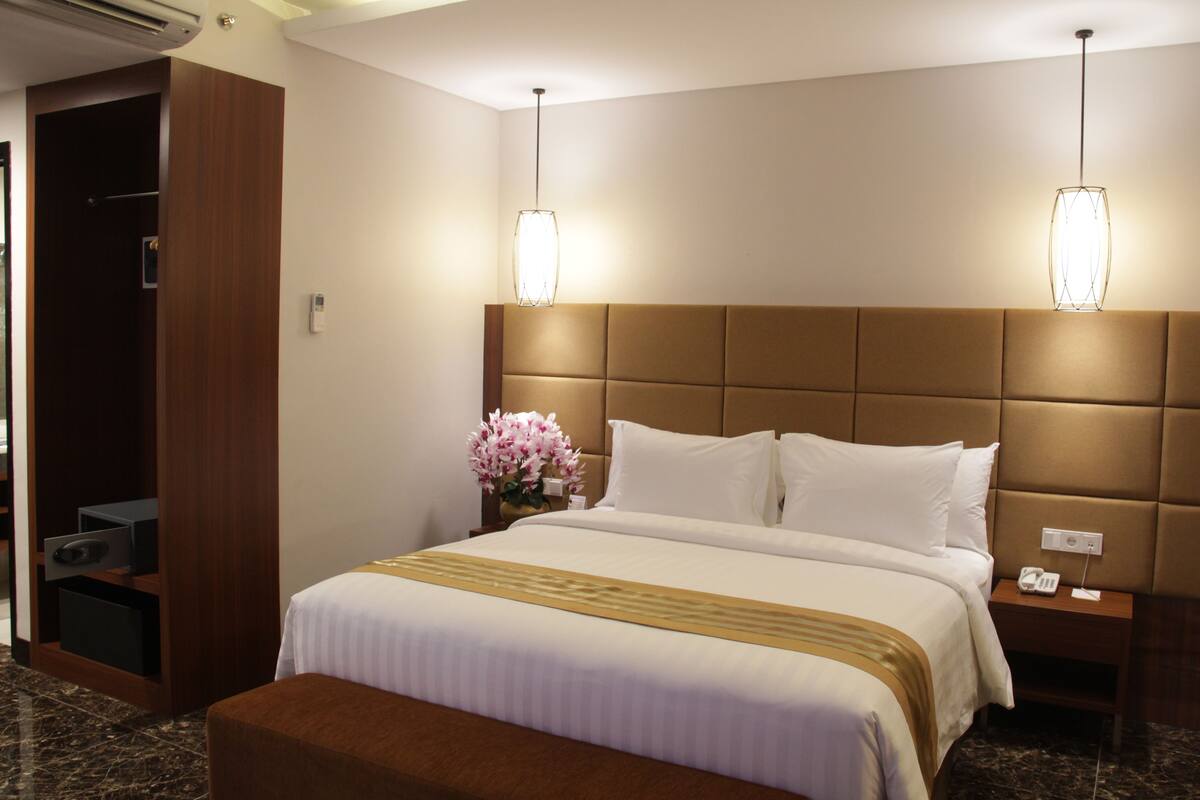 A hotel room with a large bed, a wooden wardrobe, a nightstand with a phone, and a vase of flowers.