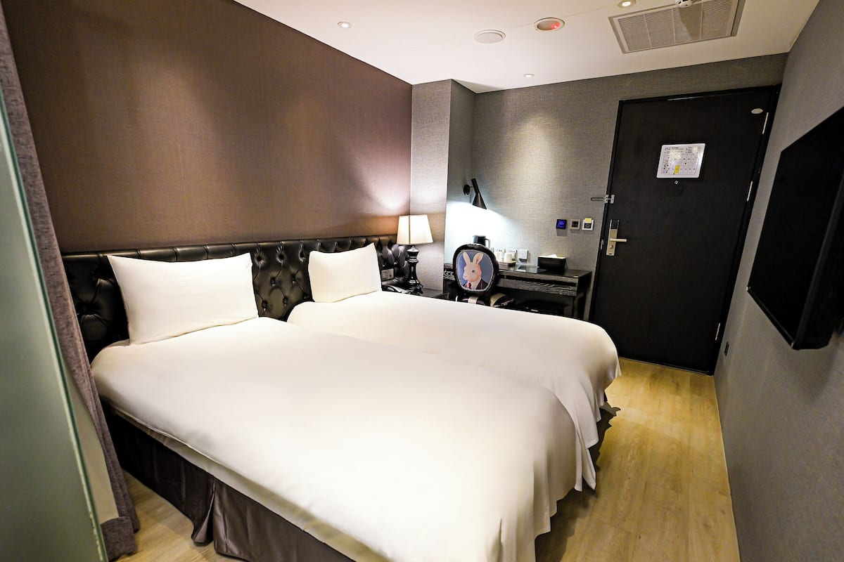 A hotel room with a large bed, a desk with a lamp, a mirror, and a door with a control panel.