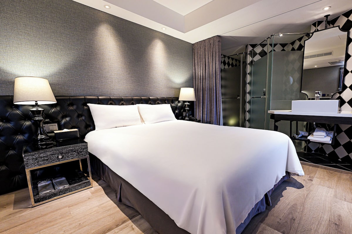 A modern hotel room with a large bed, a bedside table with a lamp, a bathroom with a glass shower, and a mirror above the sink.