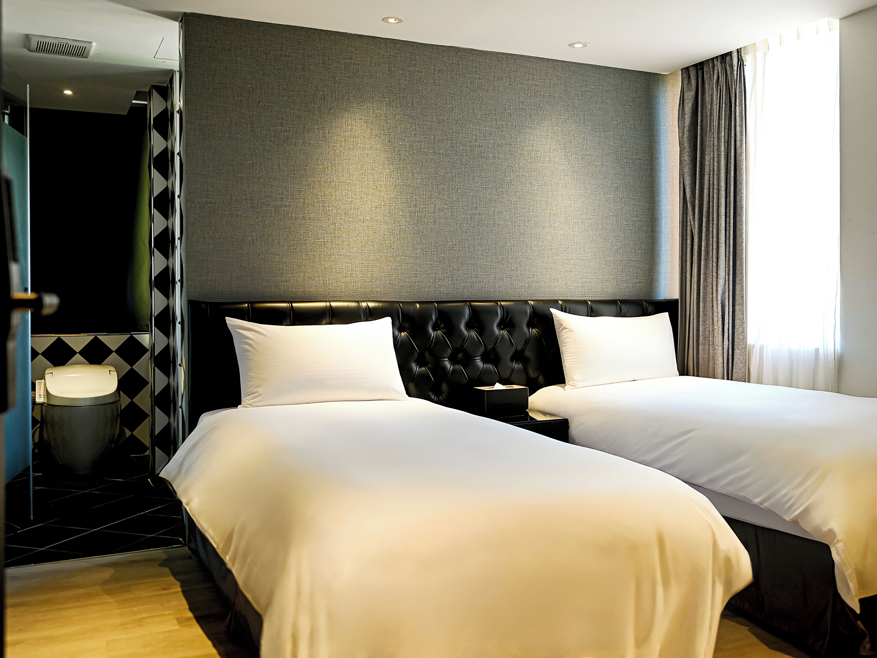 A hotel room with two beds, a dark headboard, a small bedside table, and a view of a cityscape through the window.