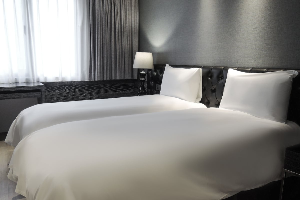 A hotel room with two beds, a dark headboard, a bedside lamp, and a window with curtains.