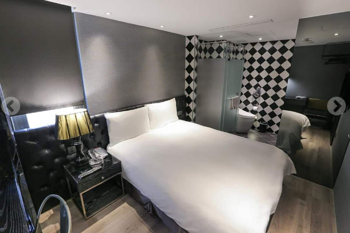 A modern hotel room with a large bed, a bedside table with a lamp, a mirror, and a black and white checkered wall.