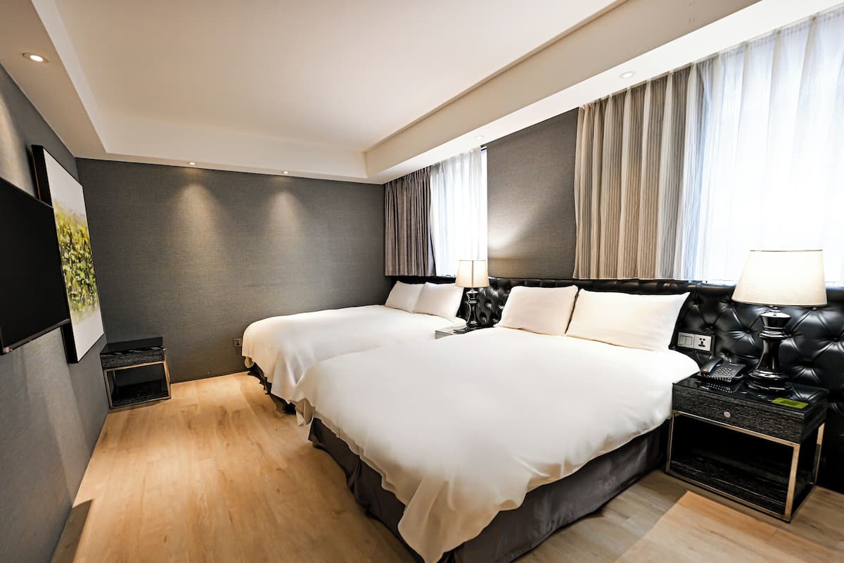 A modern hotel room with two beds, a small side table, a nightstand with a lamp, and a large window with curtains.
