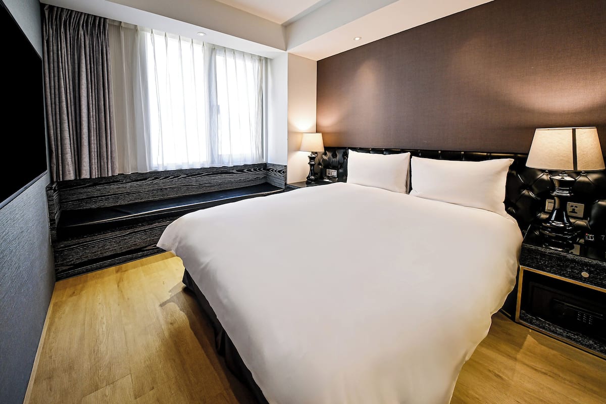 A hotel room with a large bed, a nightstand with a lamp, a window with curtains, and a dark wall.