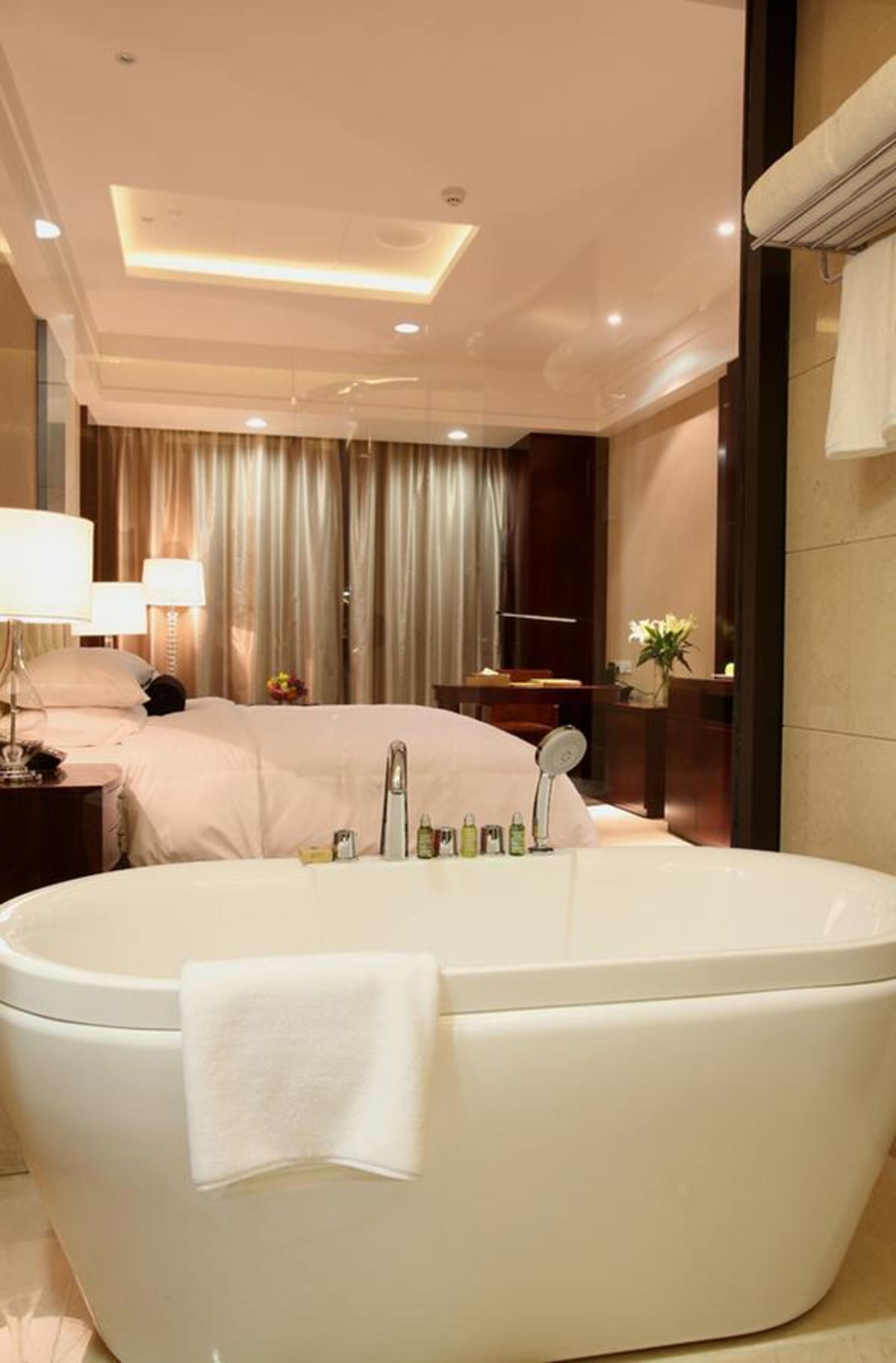 Una habitación de hotel moderna con una bañera grande, una cama con cabecero, una mesita de noche con lámpara y un espejo.