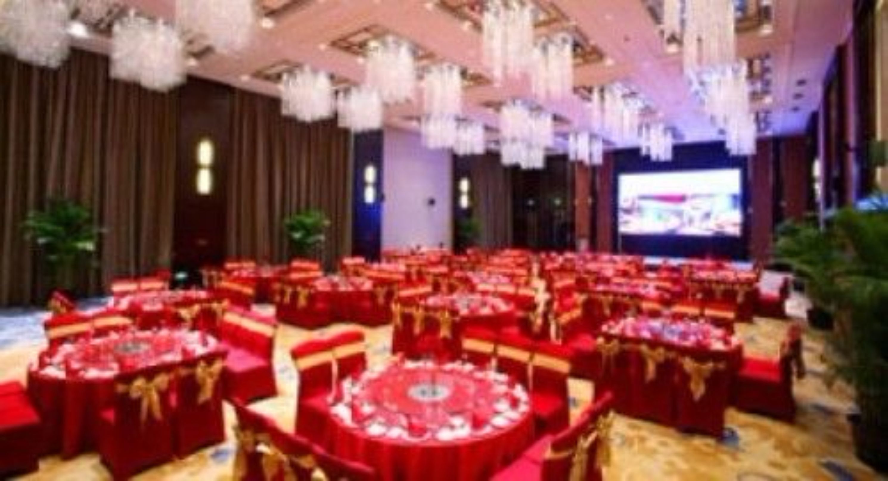 A banquet hall with round tables covered in red tablecloths and gold sashes, arranged in rows.