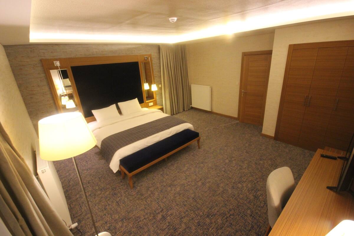 A hotel room with a large bed, a bedside table with a lamp, a desk with a chair, and a wardrobe.