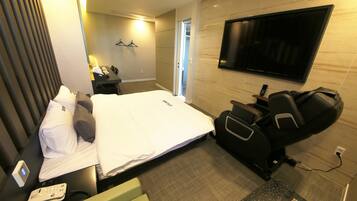 A hotel room with a bed, a massage chair, a TV, a desk with a lamp, and a small table with a remote control.