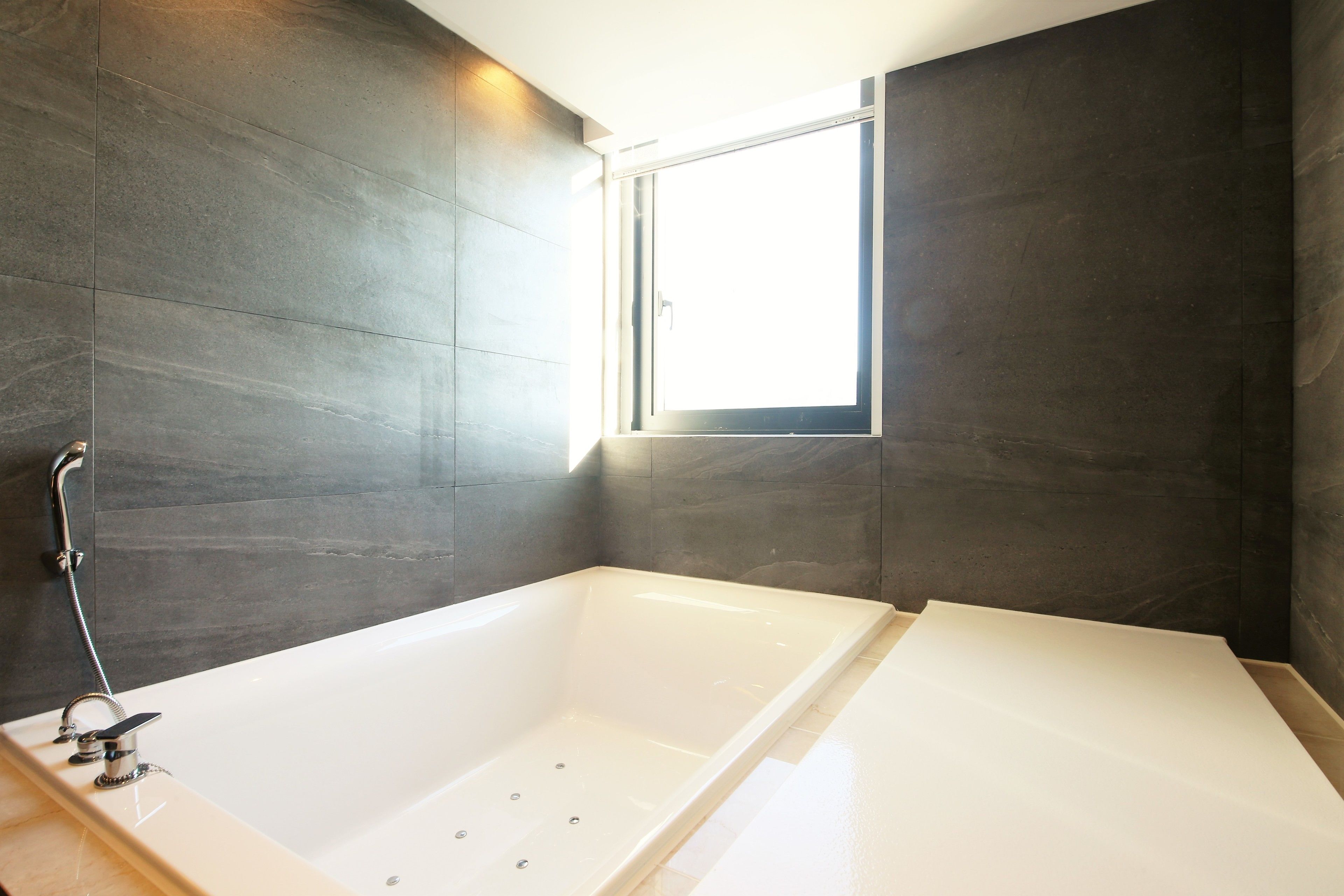 Une salle de bain moderne avec une grande baignoire, des murs recouverts de carreaux foncés et une fenêtre laissant entrer la lumière naturelle.