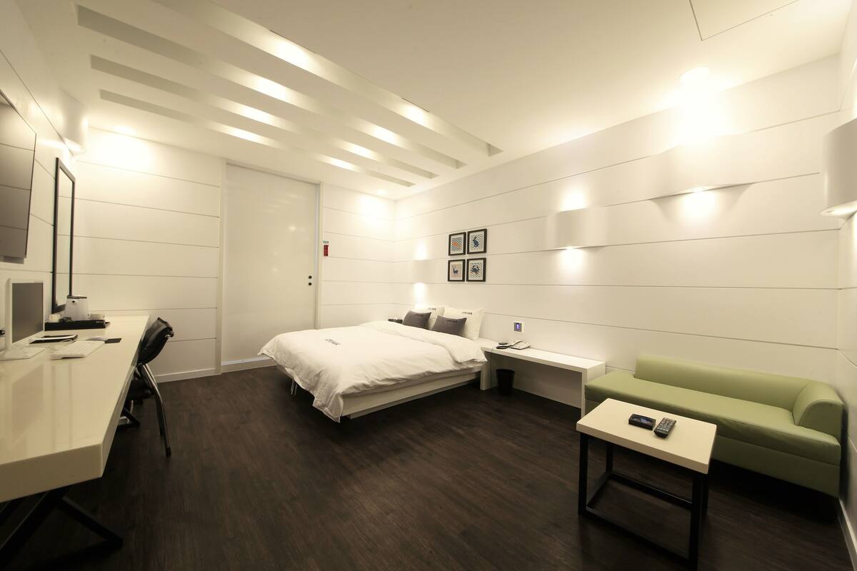 Una habitación de hotel moderna con cama, escritorio, sofá verde y una mesita con control remoto.