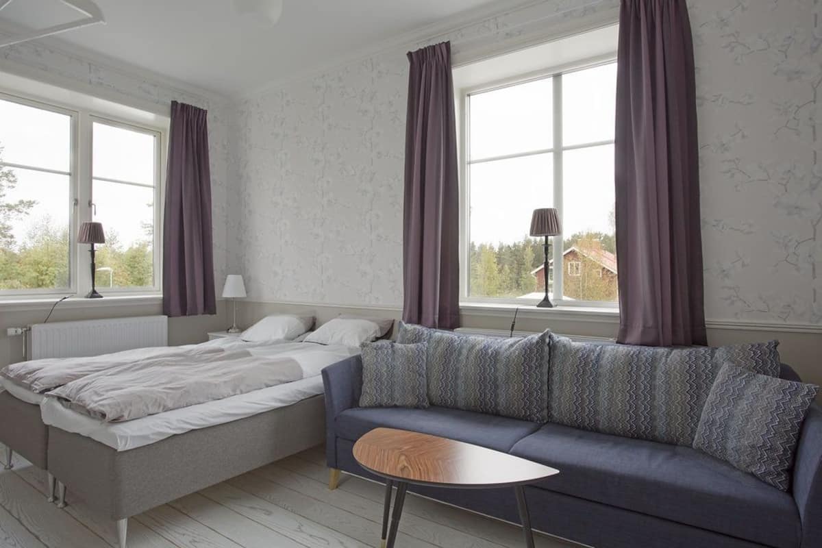 A bedroom with a bed, a sofa, a small table, and two windows with curtains.