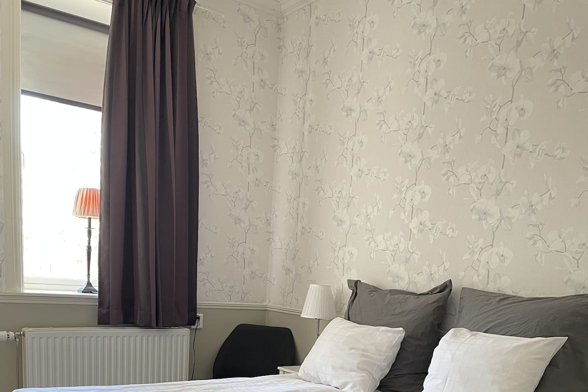 A neatly made bed with white and grey bedding, a grey upholstered headboard, and a window with a view of the outdoors.