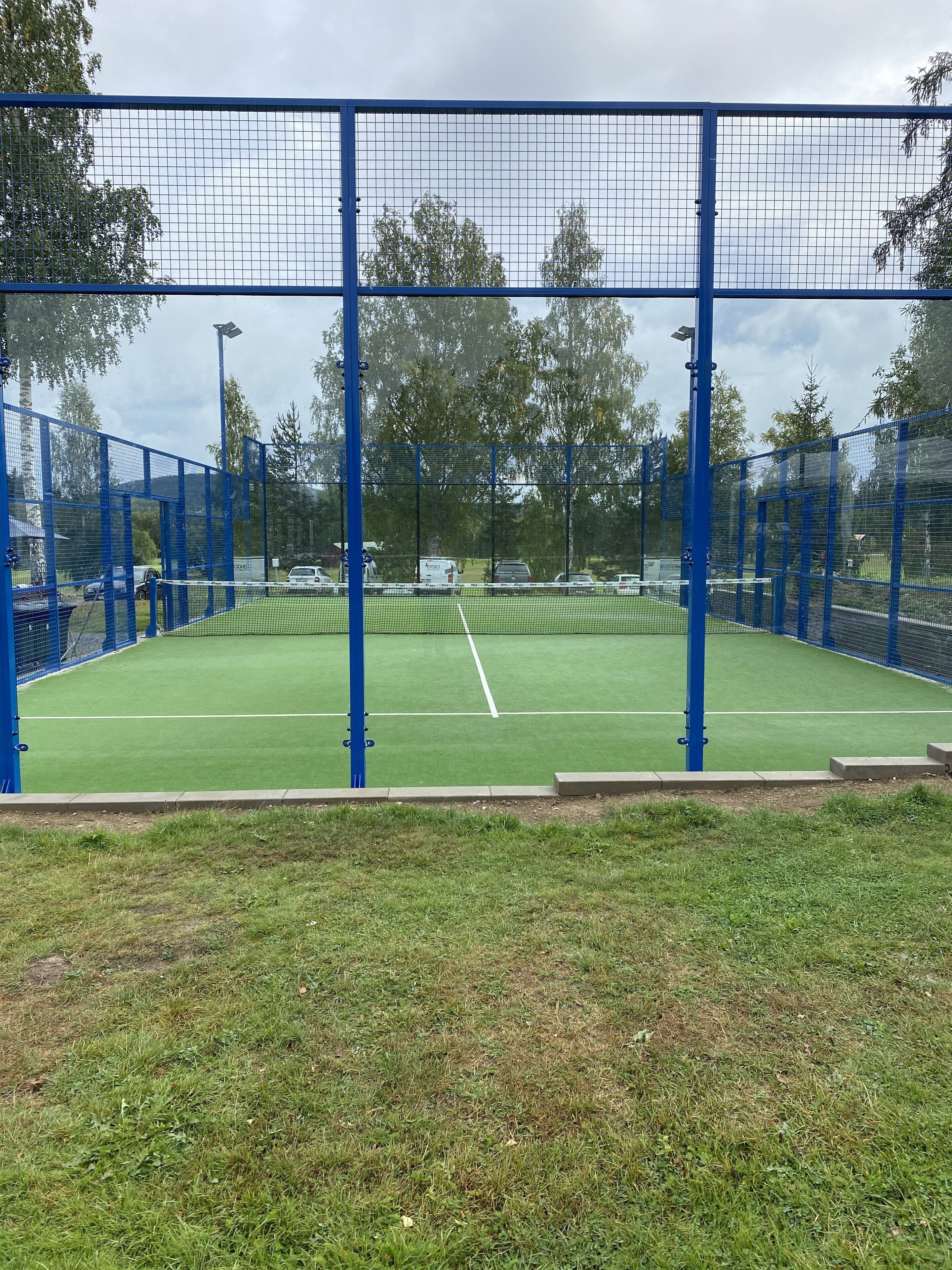 Een tennisbaan, omgeven door een blauwe hekken, met een gazon op de voorgrond.