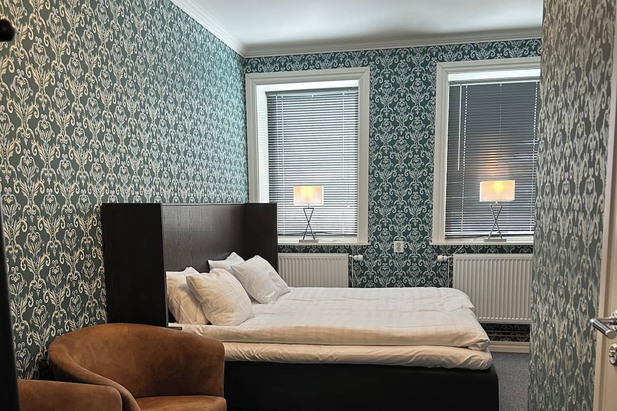 A bedroom with a bed, two bedside lamps, a radiator, and patterned wallpaper.