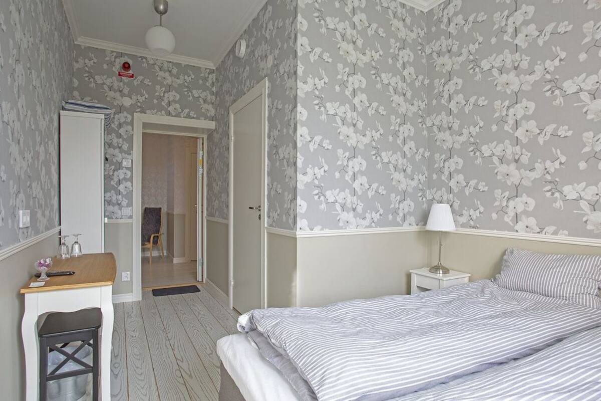 A bedroom with a bed, bedside table, and a desk with a chair. There is a door leading to another room.