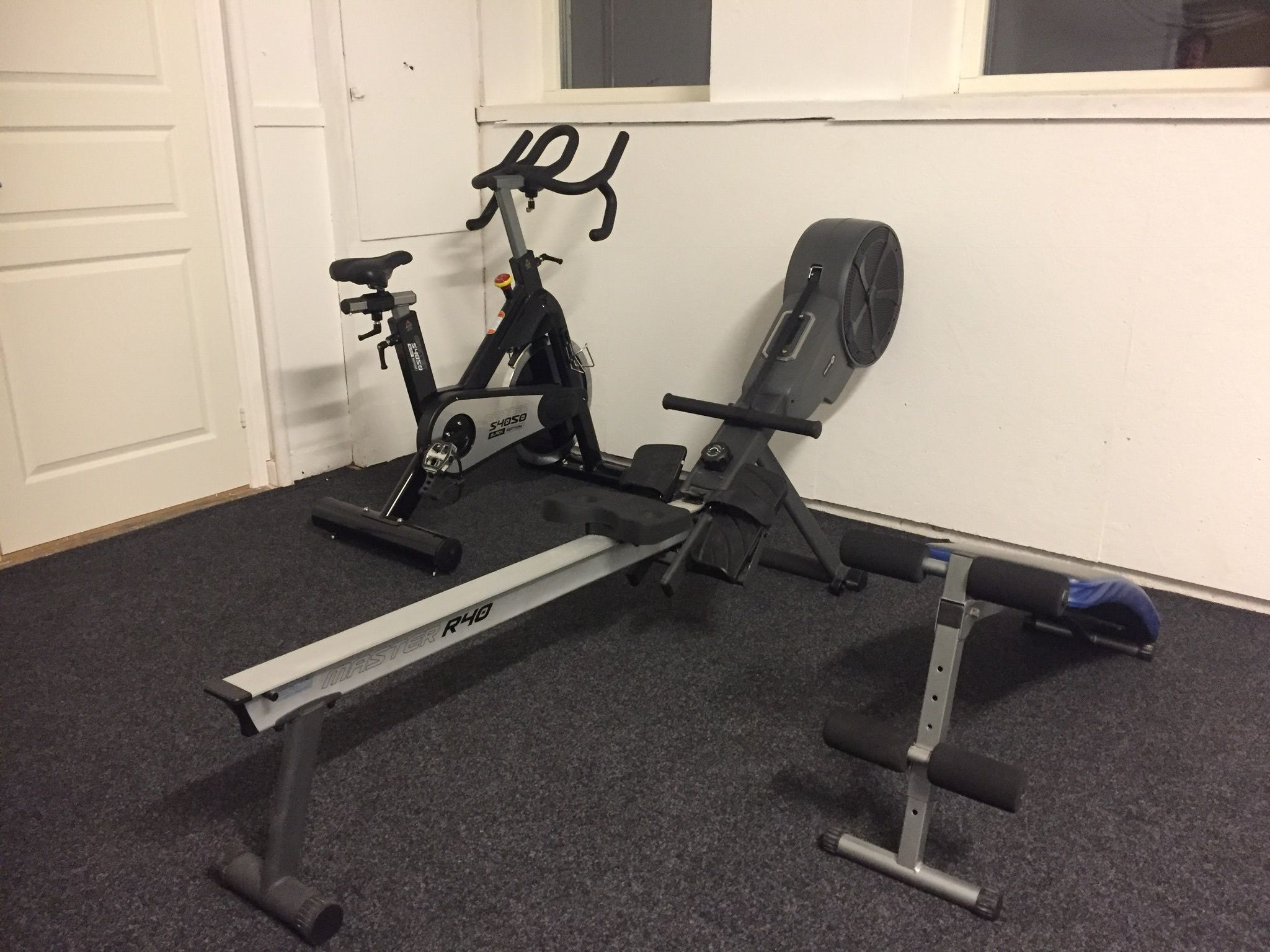 A stationary bike and a rowing machine in a room with carpeted flooring and white walls.