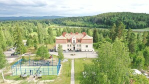 A large, two-story building with a symmetrical facade, surrounded by lush greenery and a tennis court. - Norrfly Herrgård AB (Kilafors)