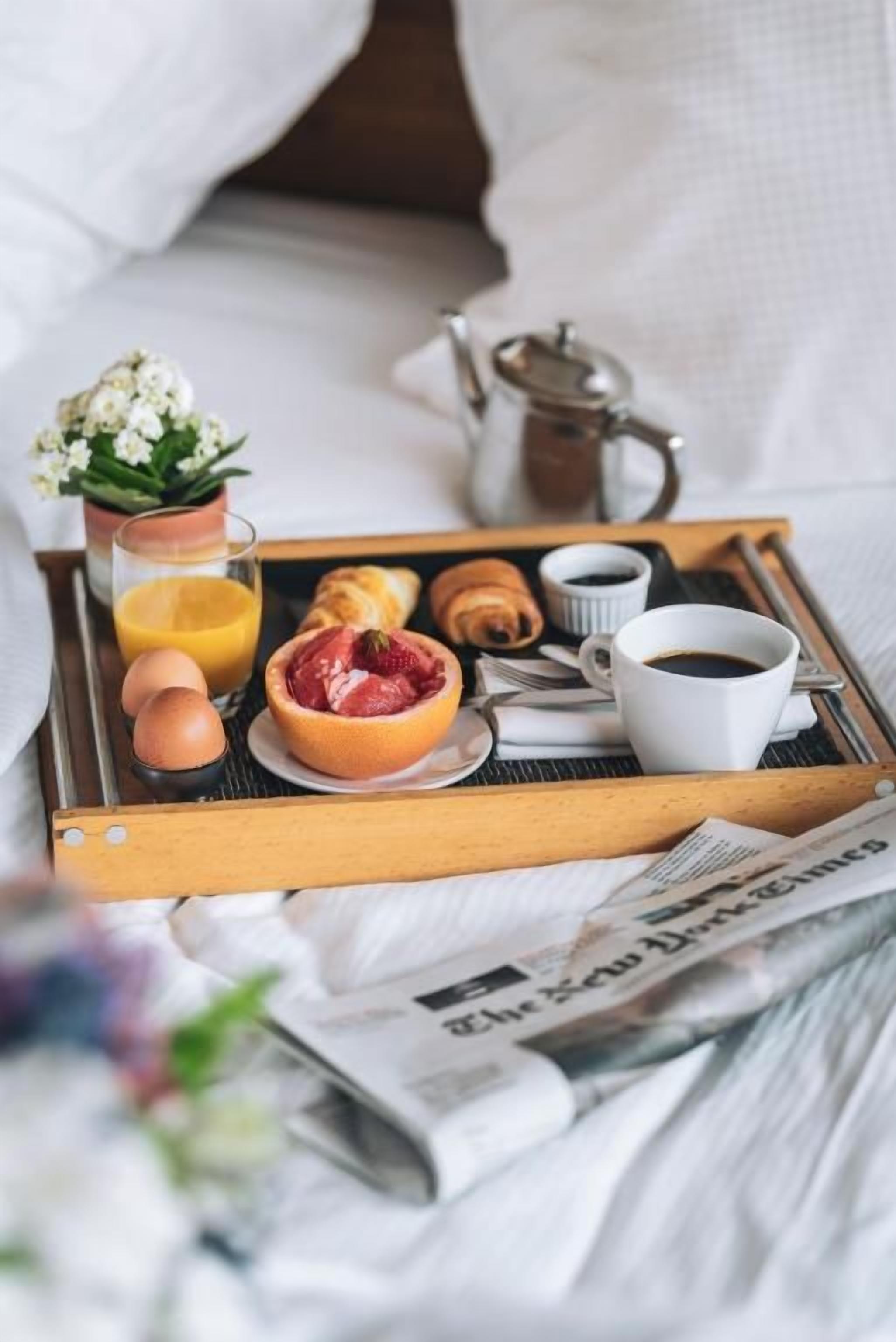 Una bandeja de desayuno con huevos, fruta, pasteles y bebidas sobre una cama con almohadas y un periódico.