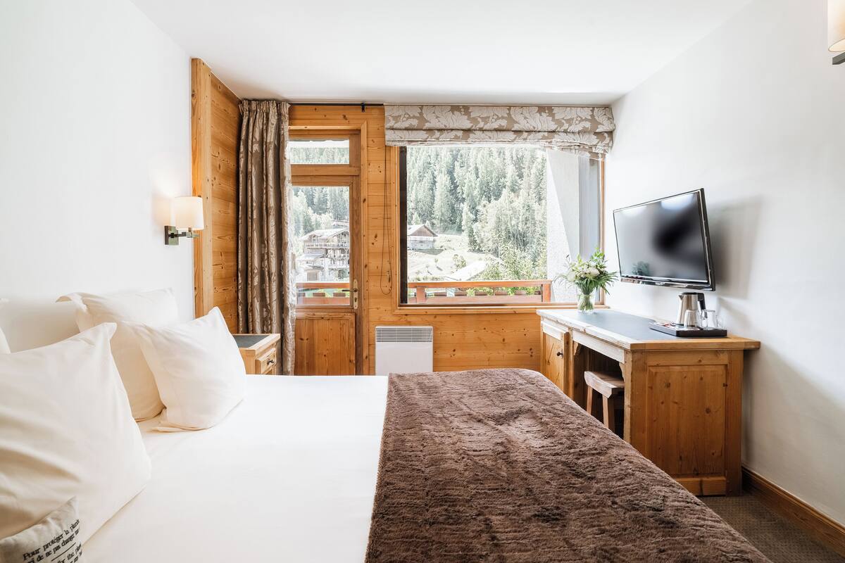 Een slaapkamer met een bed, een bureau, een televisie en een raam met uitzicht op een bos.