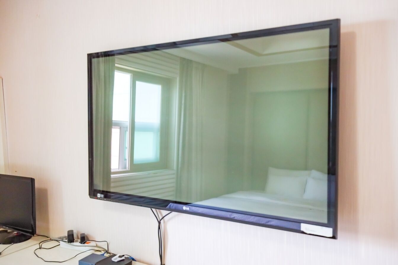 A bedroom with a large flat-screen TV mounted on the wall, a bed with white bedding, and a window with curtains.