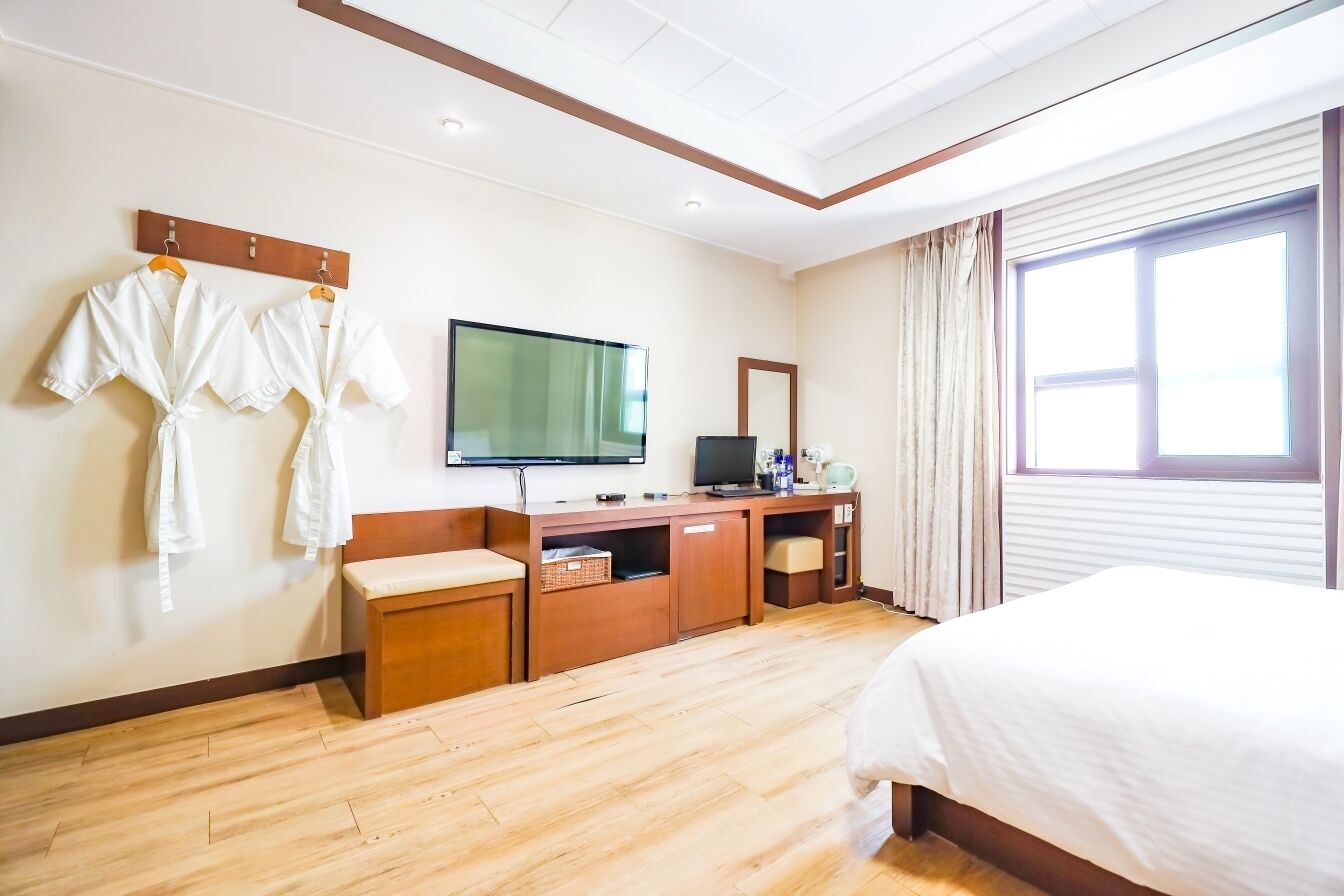 A hotel room with a bed, a TV, a wooden desk, a bench, and a wall-mounted TV.