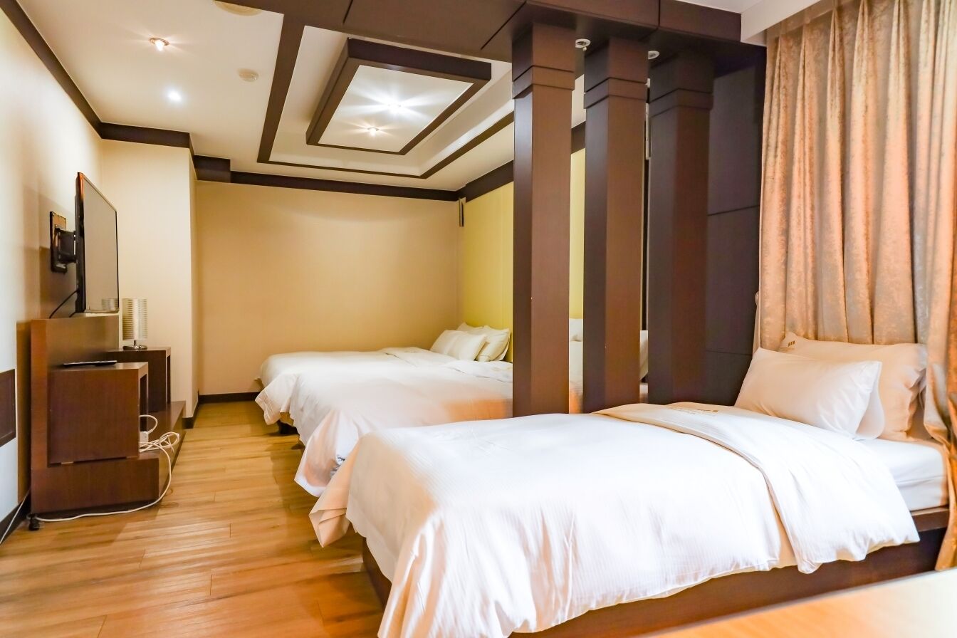 Une chambre d’hôtel avec deux lits, un plancher en bois, un luminaire au plafond et un téléviseur posé sur une commode en bois.