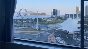 A view from a window overlooking a cityscape with a prominent Ferris wheel, modern buildings, and a body of water. - Paulownia Hostel & Guest House (Yeosu)