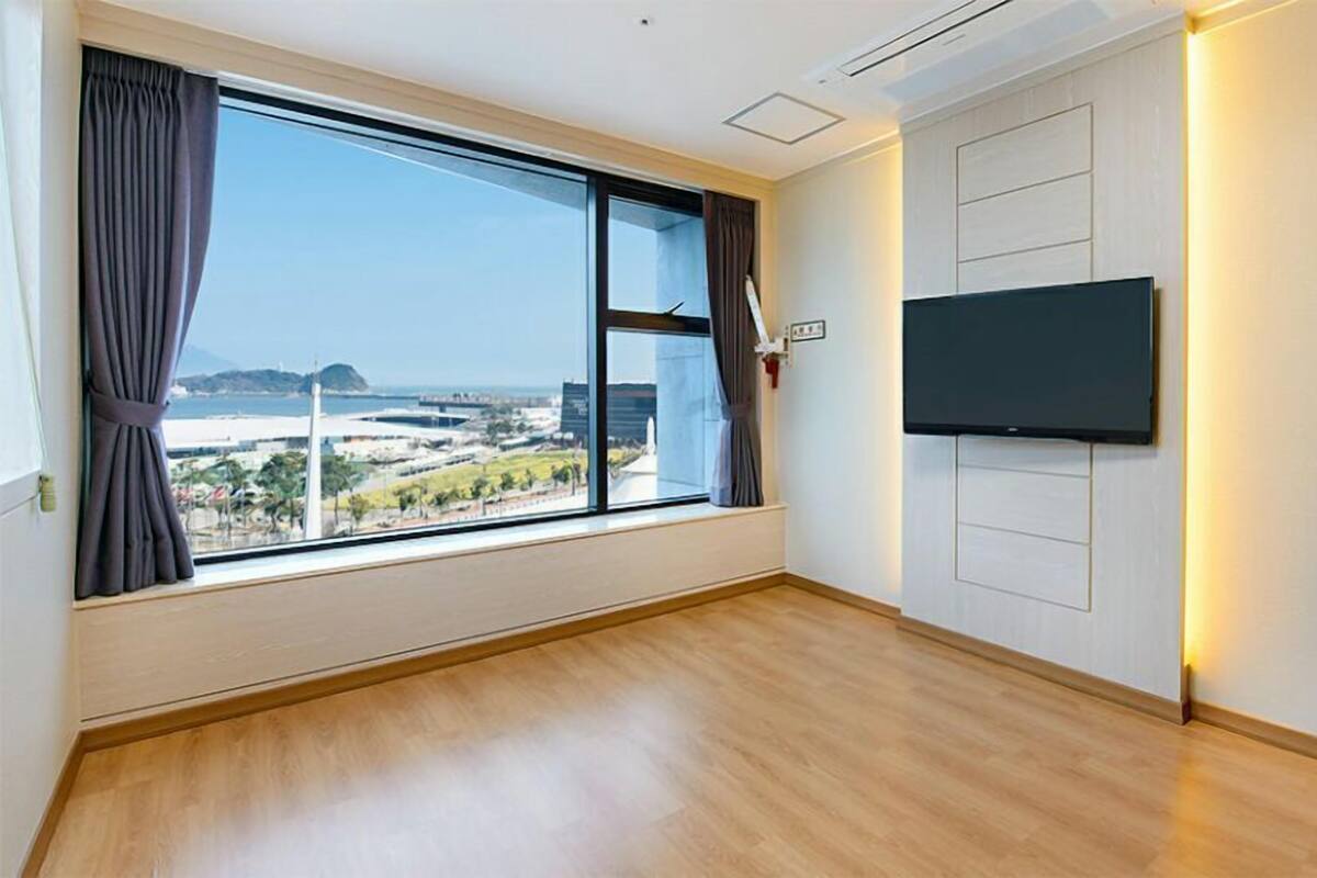 Ein Zimmer mit großem Fenster, einem an der Wand befestigten Flachbildfernseher und Holzboden.