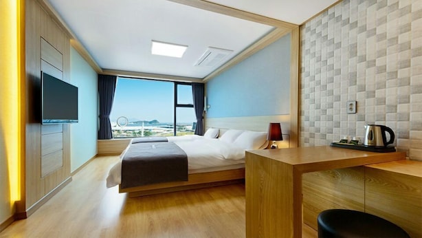 A modern hotel room with a bed, a desk, a TV, and a view of the city. - Paulownia Hostel & Guest House (Yeosu)