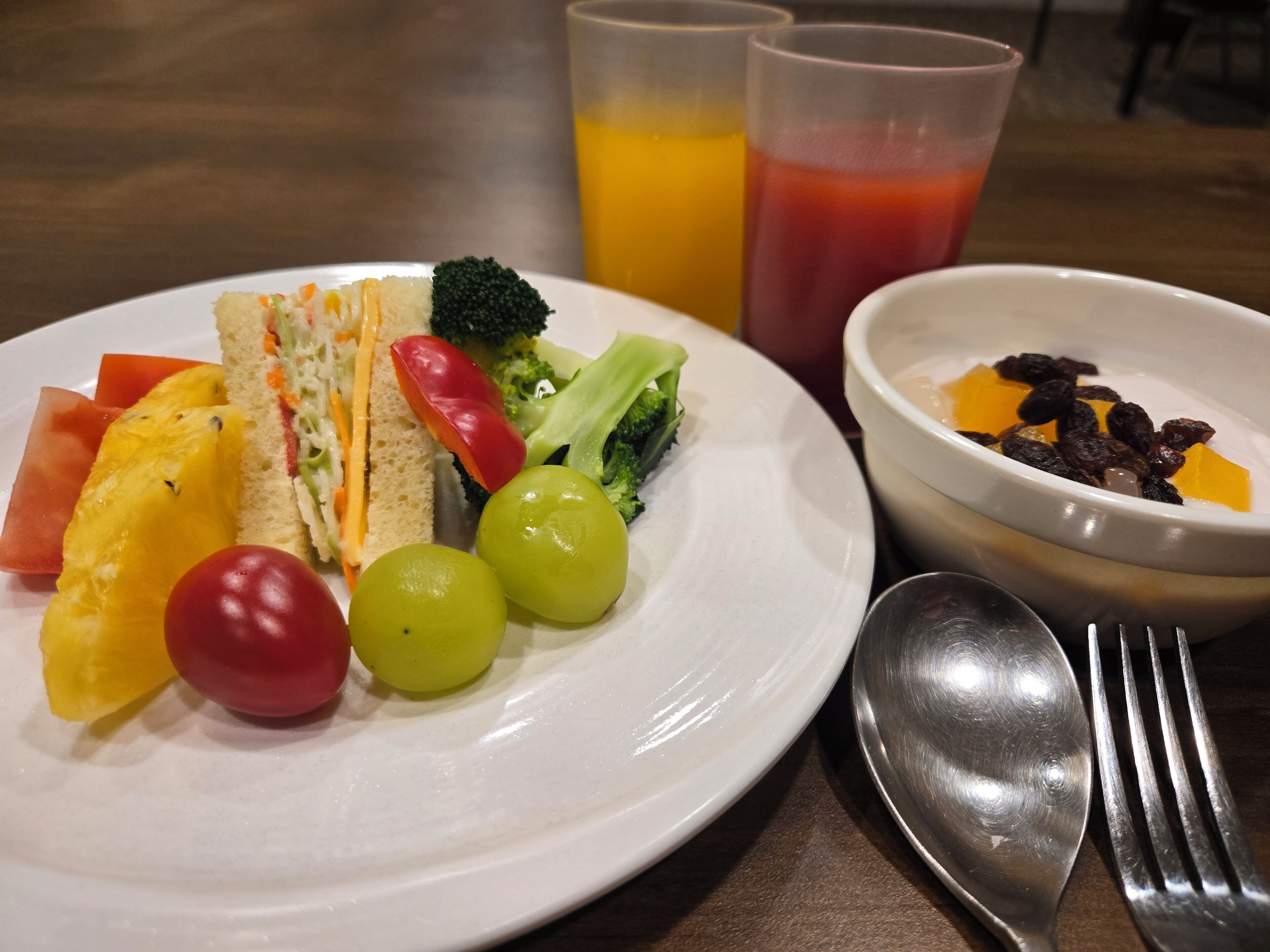 A plate with various fruits and vegetables, two glasses of juice, a bowl of cereal, and cutlery on a table.